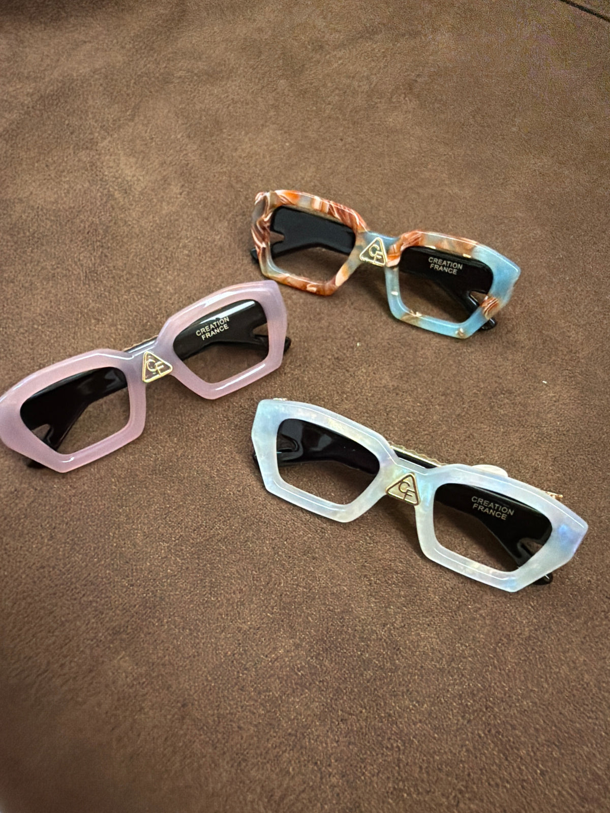 Three glasses-shaped hair clips in pink, pale pearlescent tones and a brown-and-blue mixed pattern on brown fabric.