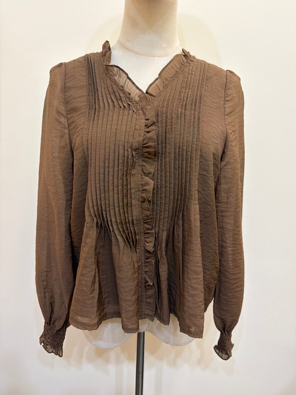 H48 brown long-sleeve blouse on a mannequin, with a ruffle-trim neckline, vertical pintuck front and smocked cuffs.
