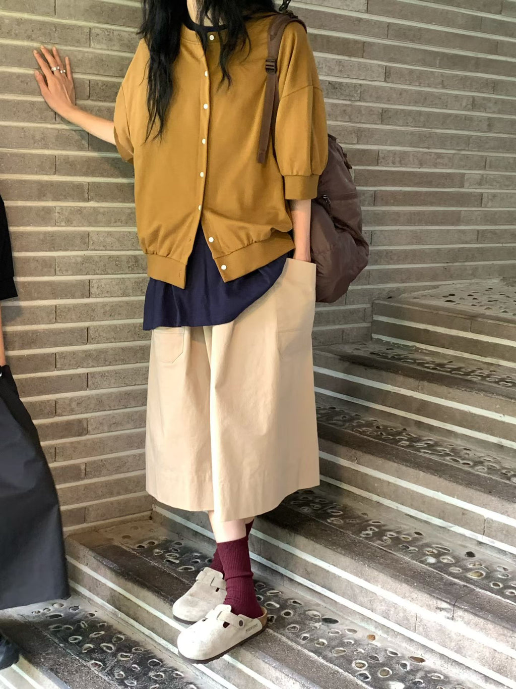 H62 brown jacket with the front mostly buttoned, layered over a navy top and beige wide-leg bottoms beside a staircase.