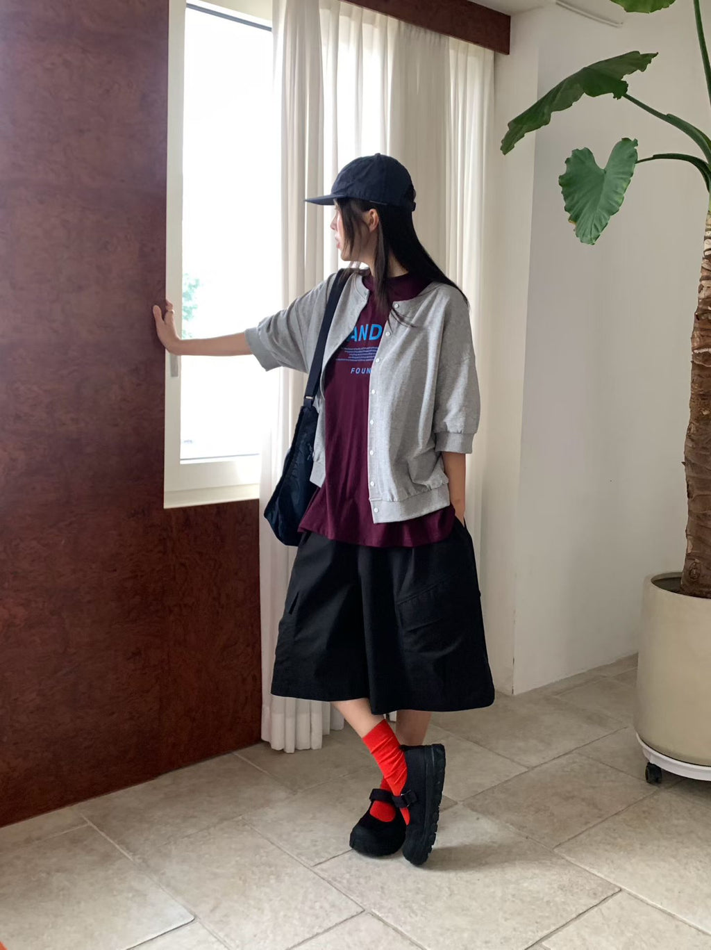 Full-outfit view of the H62 gray jacket worn open with a burgundy T-shirt, black shorts, red socks and black shoes by a window.