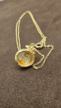 Close-up of a gold-tone round pendant with a seated Buddha figure, pale background and dotted border beside its fine chain.