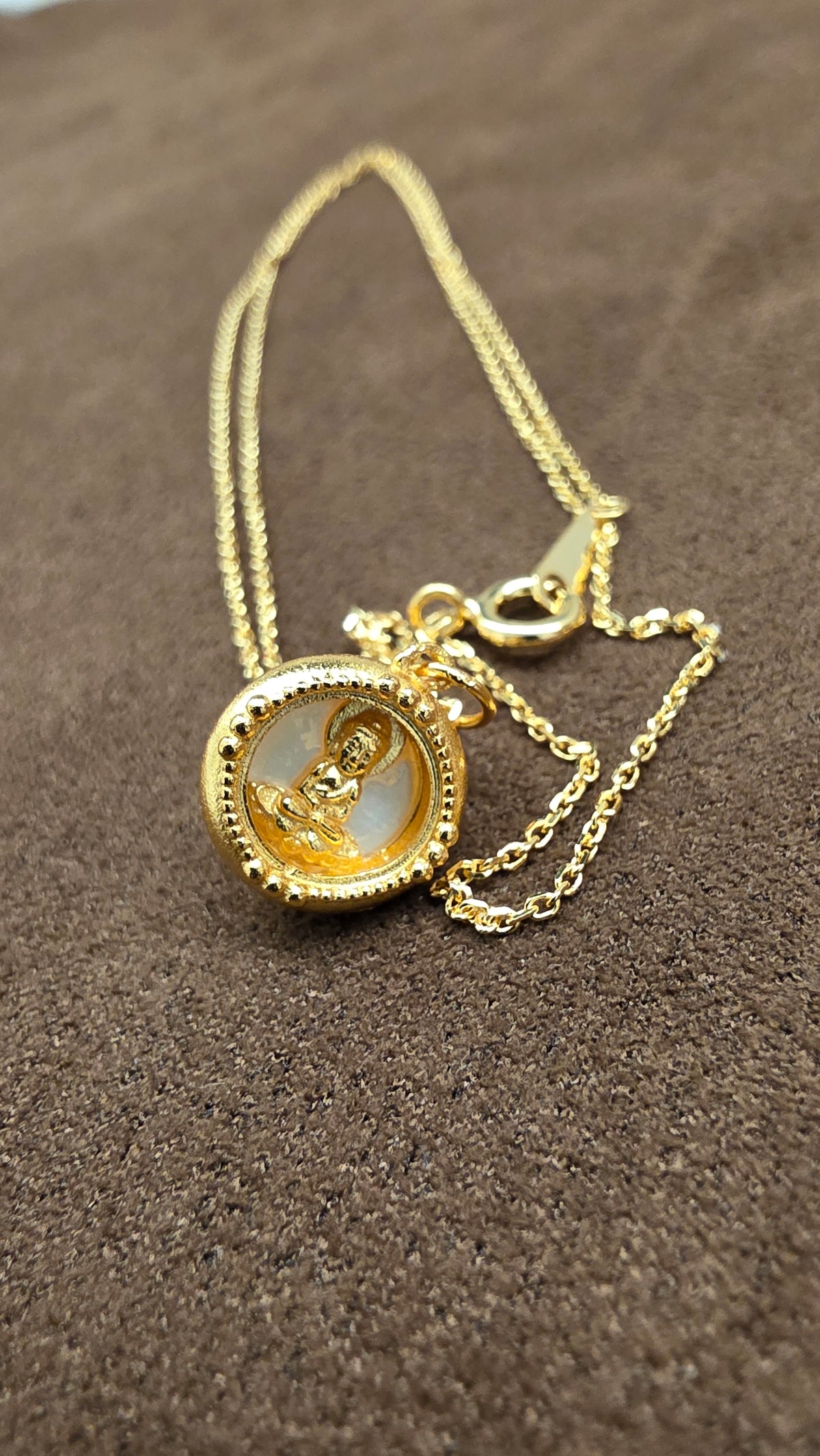 Close-up of a gold-tone round pendant with a seated Buddha figure, pale background and dotted border beside its fine chain.