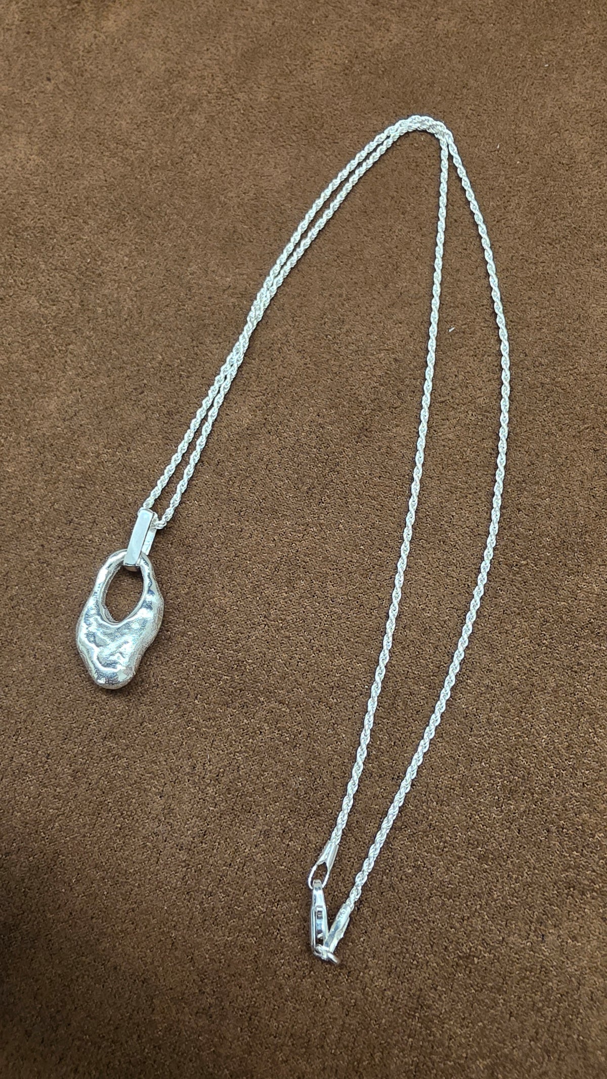A62 silver necklace with an irregular textured open pendant, rectangular bail and fine rope-like chain on brown fabric.