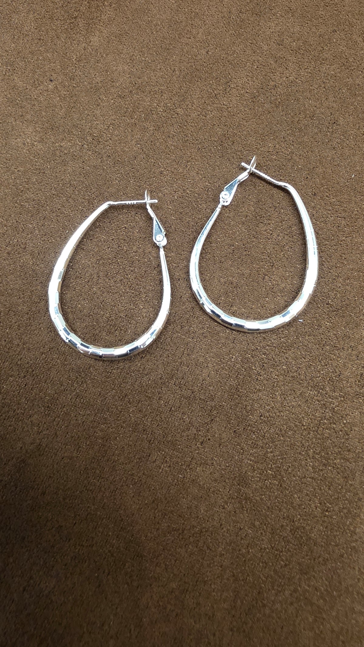 Pair of silver teardrop-shaped hoop earrings with small reflective surface markings and upper post clasps on brown fabric.