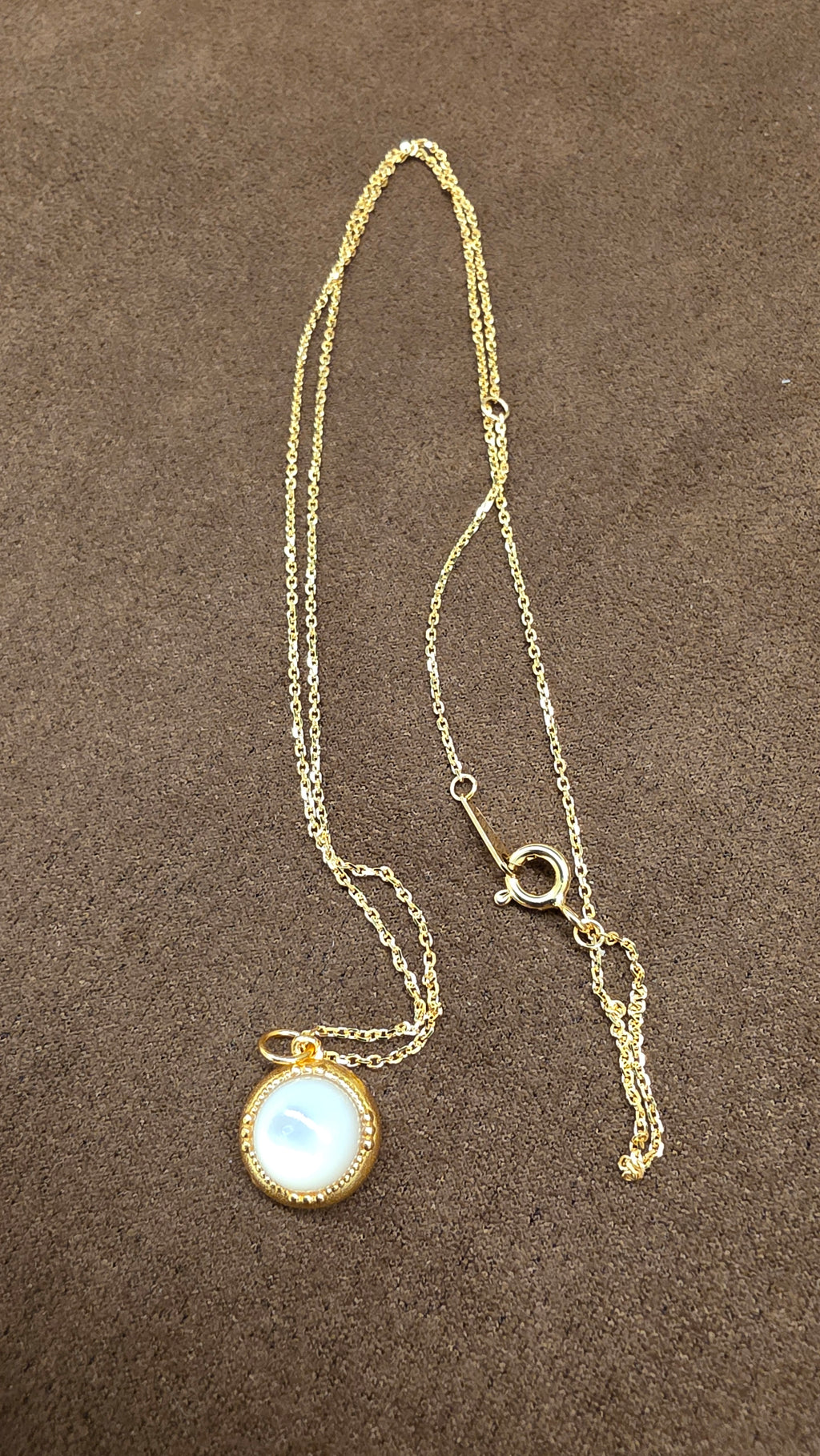 Reverse view of a round gold-tone pendant with a pale reflective centre, fine chain and circular clasp on brown fabric.