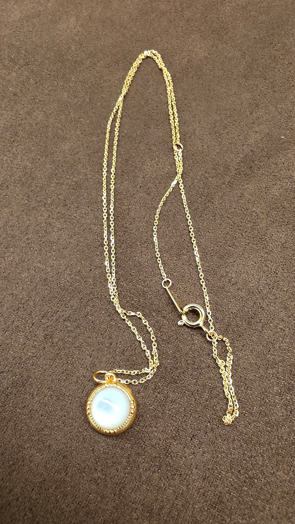 Reverse view of a round gold-tone pendant with a pale reflective centre, fine chain and circular clasp on brown fabric.