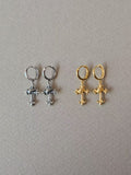 Silver-tone and gold-tone hoop earrings with ornate cross-shaped drops and diagonal wrap details, shown side by side on grey fabric.