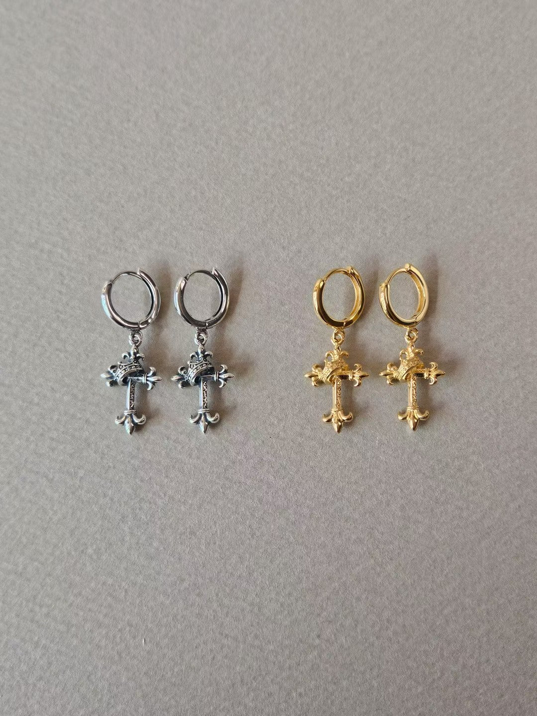 Silver-tone and gold-tone hoop earrings with ornate cross-shaped drops and diagonal wrap details, shown side by side on grey fabric.