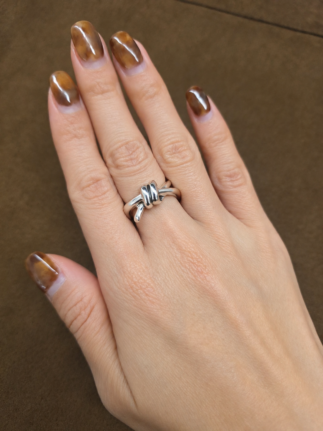 A63 silver-tone ring with a raised double-loop knot and overlapping curved band, worn on a hand against brown fabric.