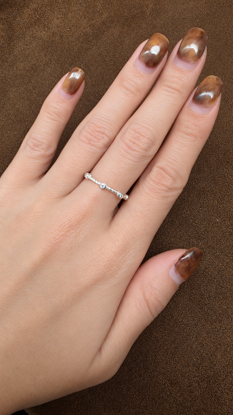 Silver-tone slender twisted band ring with small round clear-looking accents, worn on a hand against brown fabric.